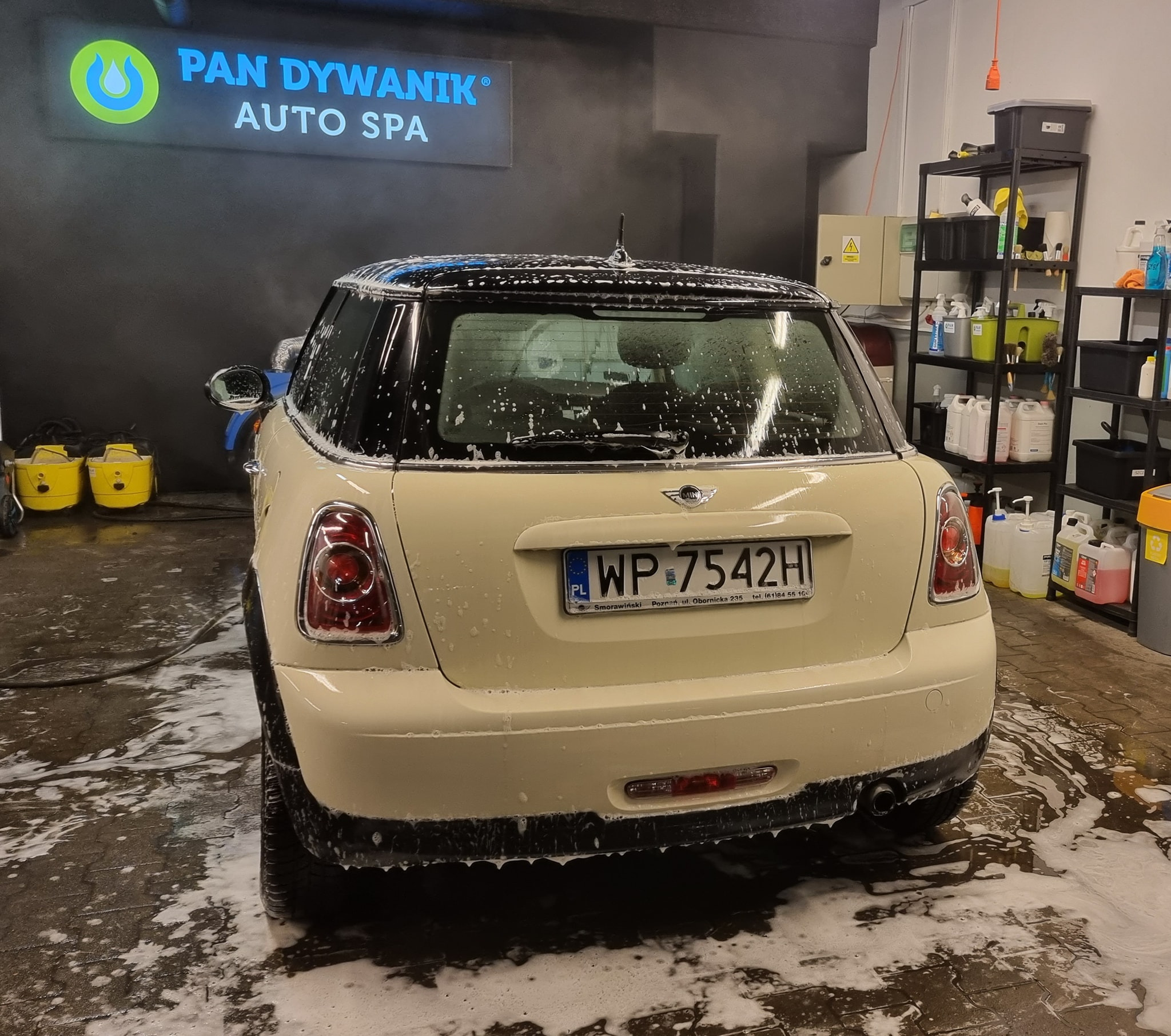 Kremowy Mini Cooper w trakcie mycia w myjni samochodowej, pokryty pianą, widoczny tył pojazdu i logo firmy 'Pan Dywanik Auto Spa' w tle.