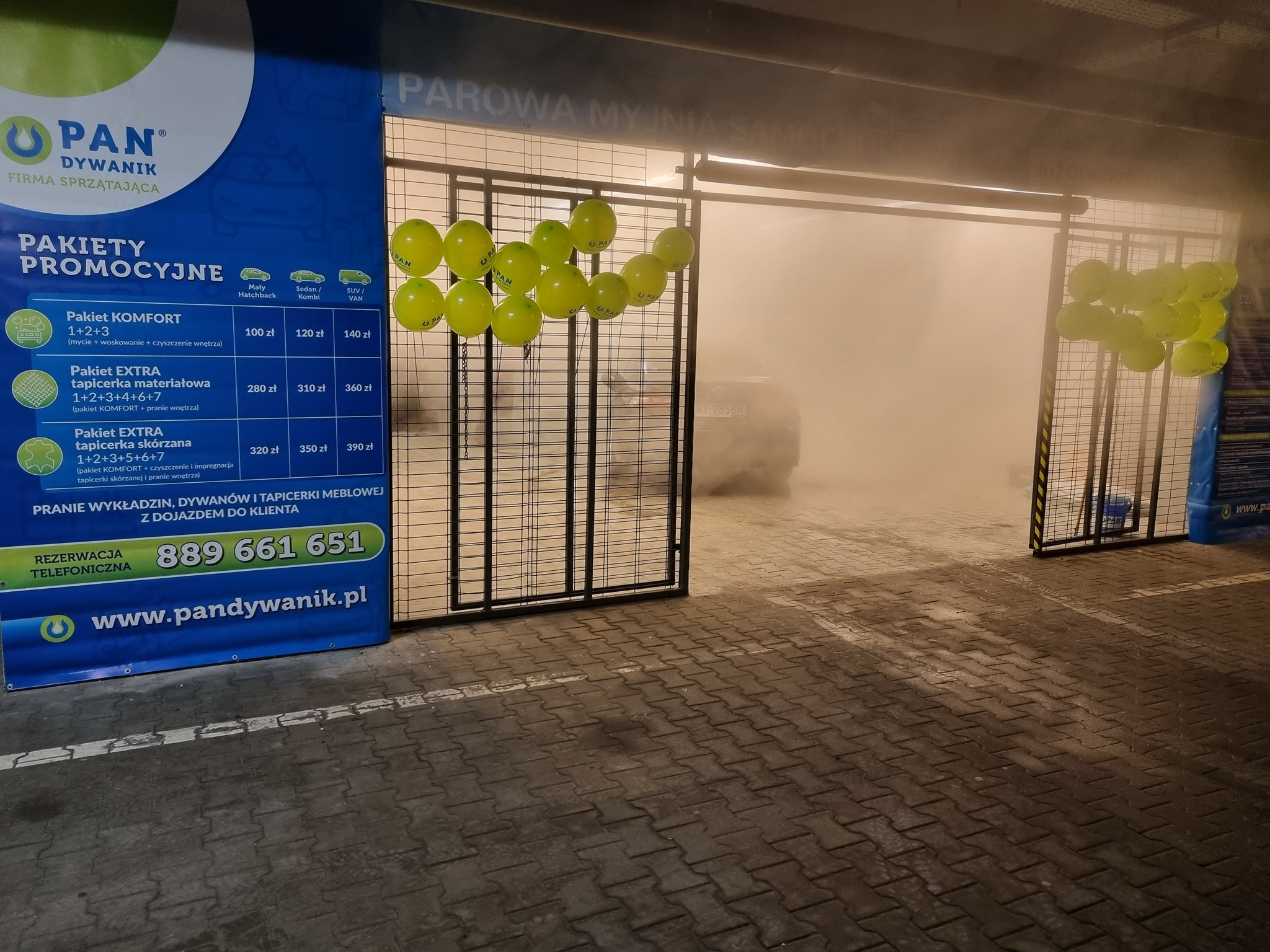 Wjazd do parowej myjni samochodowej, ozdobiony żółtymi balonami, z widocznym samochodem wewnątrz i banerem reklamowym pakietów usług po lewej stronie.