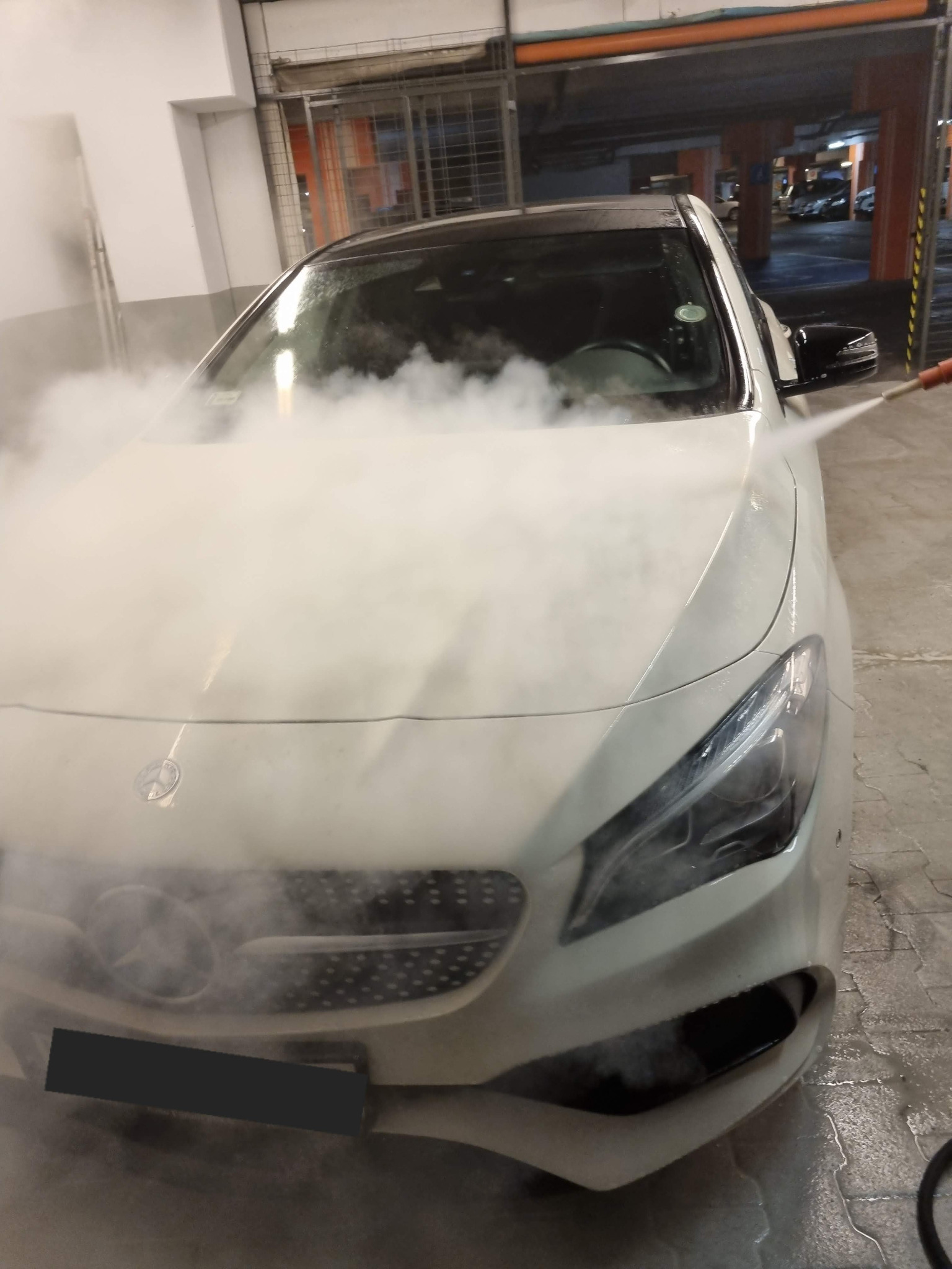 Biały samochód marki Mercedes czyszczony parą wodną pod ciśnieniem w myjni samochodowej, widok z przodu na maskę i reflektory, w tle parking wielopoziomowy.