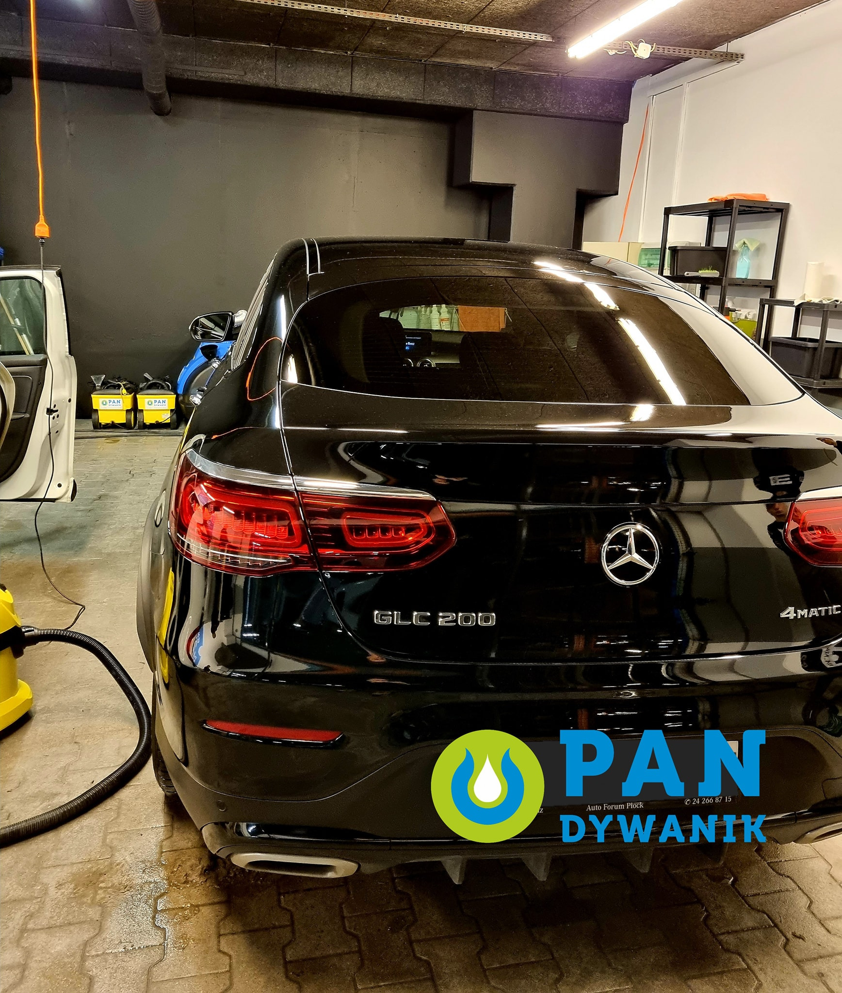 Czarny Mercedes GLC 200 w myjni, widoczne odkurzacze piorące, mokra posadzka z kostki brukowej, logo firmy nałożone na zdjęcie.