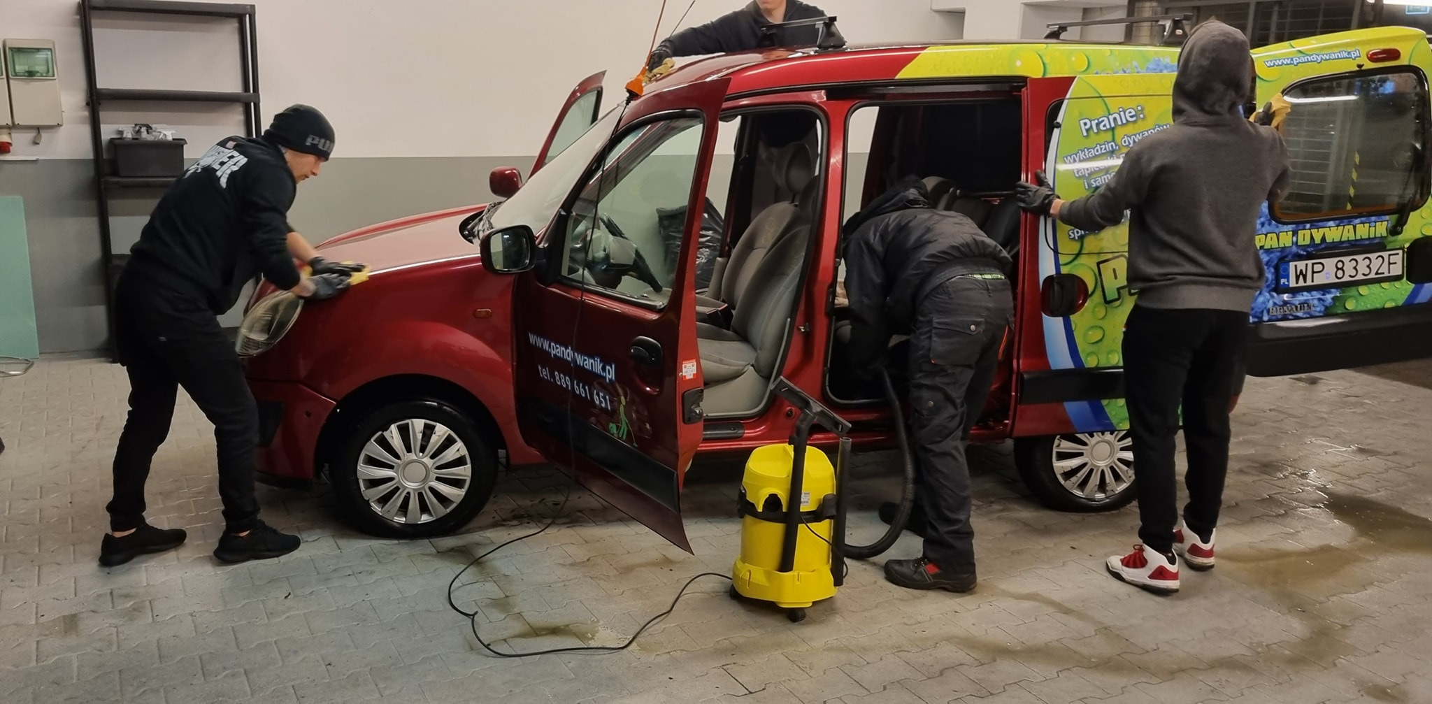Trzech mężczyzn czyści wnętrze i karoserię czerwonego samochodu dostawczego z reklamą firmy, używając odkurzacza i gąbek, widoczny żółty odkurzacz przemysłowy.