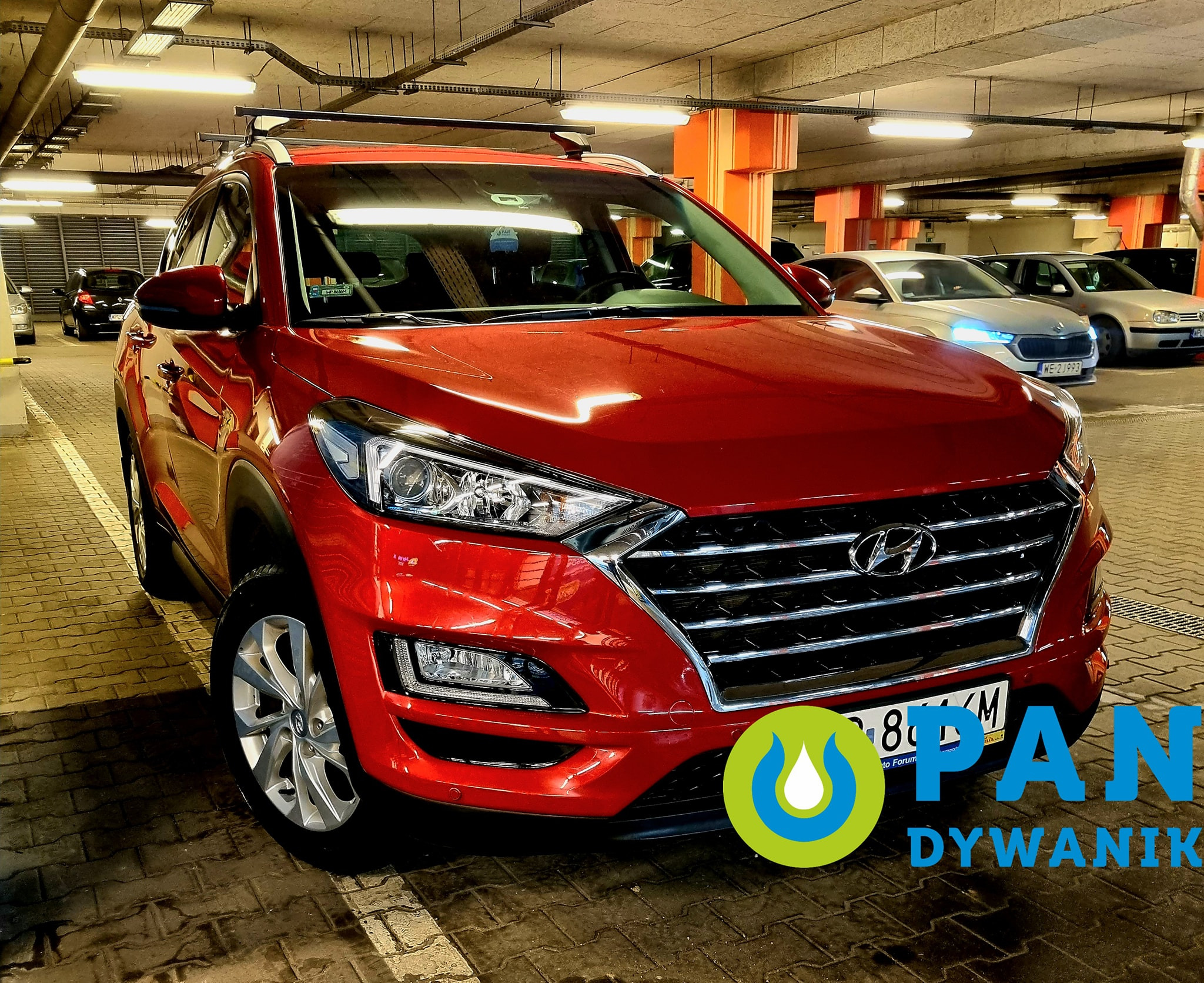 Czerwony samochód SUV marki Hyundai na parkingu podziemnym, z nałożonym logo firmy zajmującej się czyszczeniem dywaników samochodowych.