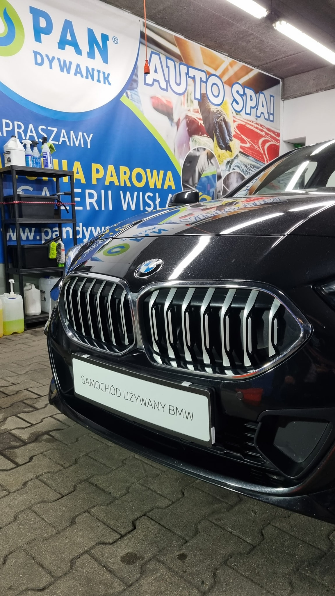 Czarny samochód BMW z widocznym grillem i tablicą 'Samochód używany BMW' na tle baneru reklamowego myjni samochodowej z napisem 'Auto Spa'.