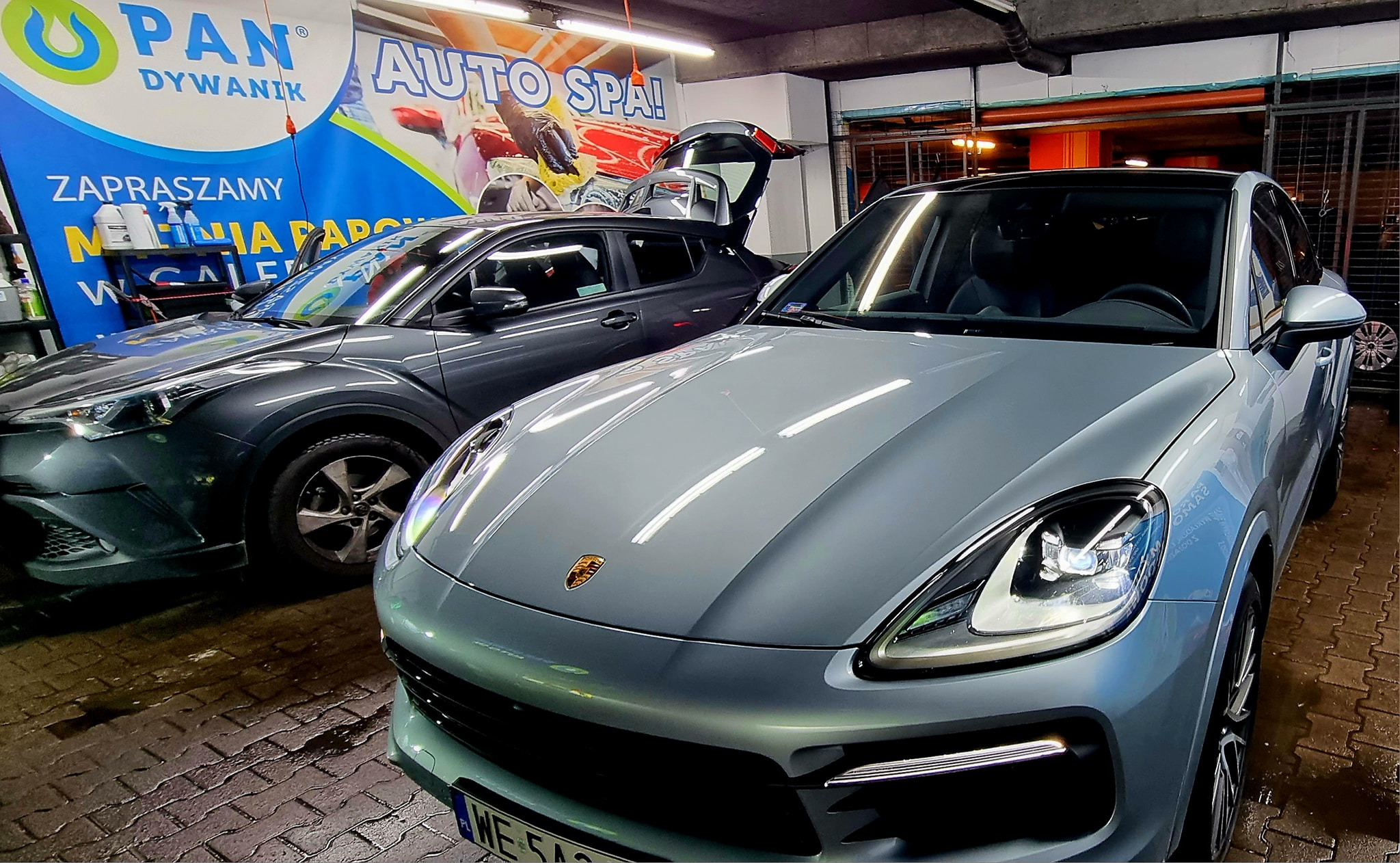 Dwa samochody, szary Toyota CHR z otwartym bagażnikiem i jasnoszary Porsche Cayenne, stoją w myjni samochodowej z banerami reklamowymi w tle.