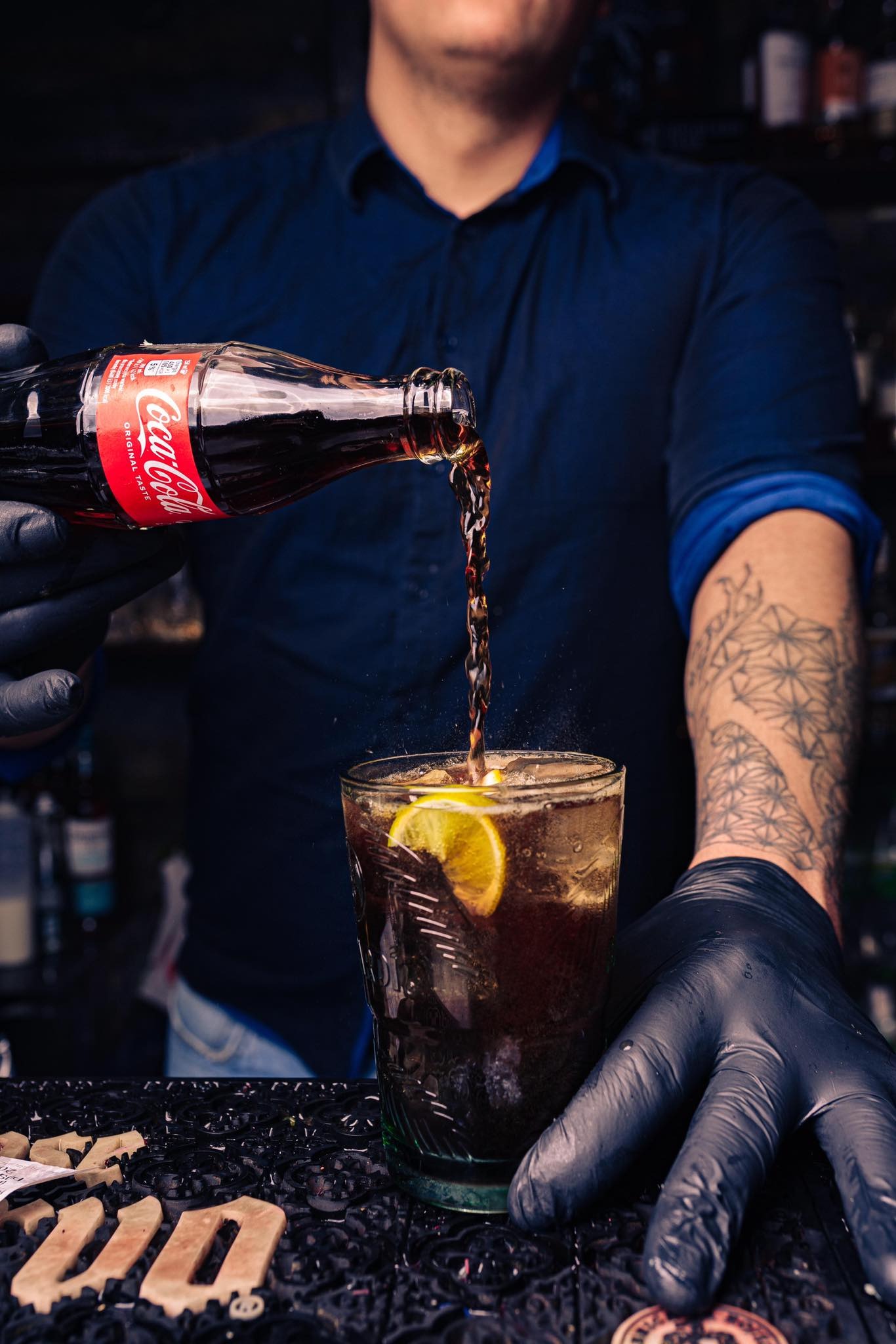 Barman w czarnych rękawiczkach nalewa napój Coca-Cola z butelki do szklanki z lodem i plasterkiem cytryny, na barze widoczne dekoracje.