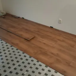 Fragment pokoju w trakcie montażu paneli podłogowych z widoczną podkładką izolacyjną, młotkiem i listwami przypodłogowymi.