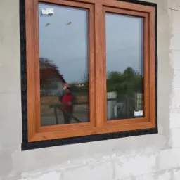 Zamontowane okno PCV o drewnopodobnej ramie w surowej ścianie z betonu komórkowego, zabezpieczone czarną taśmą uszczelniającą, z odbiciem osoby w szybie.