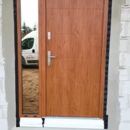 Montaż zewnętrznych drzwi wejściowych z bocznym panelem lustrzanym w surowym stanie budynku, widoczne zabezpieczenia taśmą uszczelniającą i tymczasowe drewniane podesty.