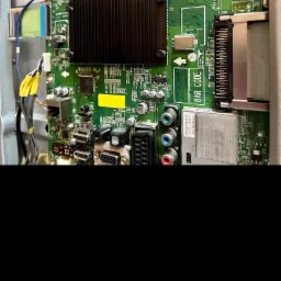 Wnętrze telewizora z widoczną płytą główną, licznymi komponentami elektronicznymi, złączami i przewodami. Widoczny radiator na układzie scalonym oraz etykieta z kodem kreskowym. Detale płyty...