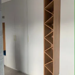 Nowoczesna szafa wnękowa z szarymi frontami i złotymi uchwytami, uzupełniona o drewniany regał na wino z trójkątnymi przegródkami.