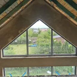 Wnętrze poddasza z surowymi ścianami, widoczne okno trójkątne z widokiem na ogród, drewniana konstrukcja dachu z zielonymi belkami.