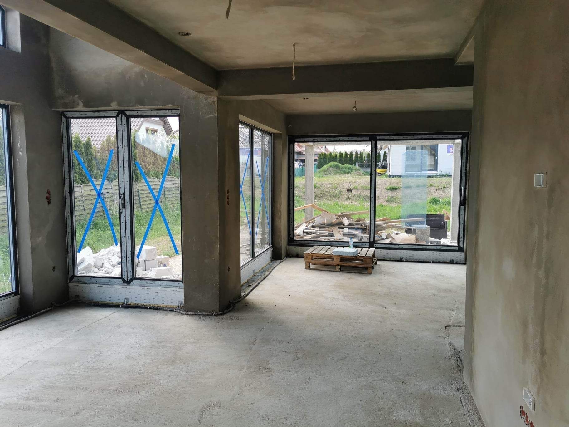 Surowy stan wnętrza domu z oknami zabezpieczonymi niebieską taśmą, widoczne ślady po pracach budowlanych na ścianach i podłodze, paleta z resztkami materiałów budowlanych.