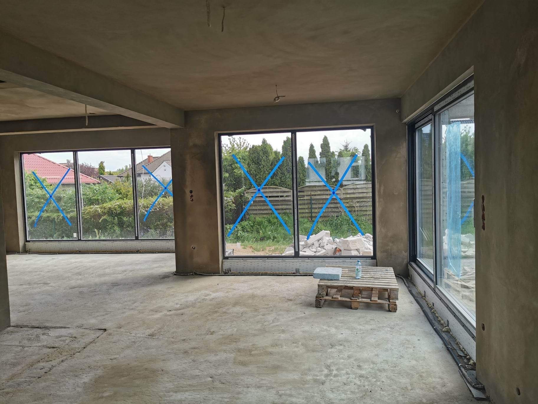 Surowe wnętrze z widocznymi świeżo wykonanymi ścianami i sufitem, zabezpieczone okna folią i taśmą, paleta z wodą na podłodze.