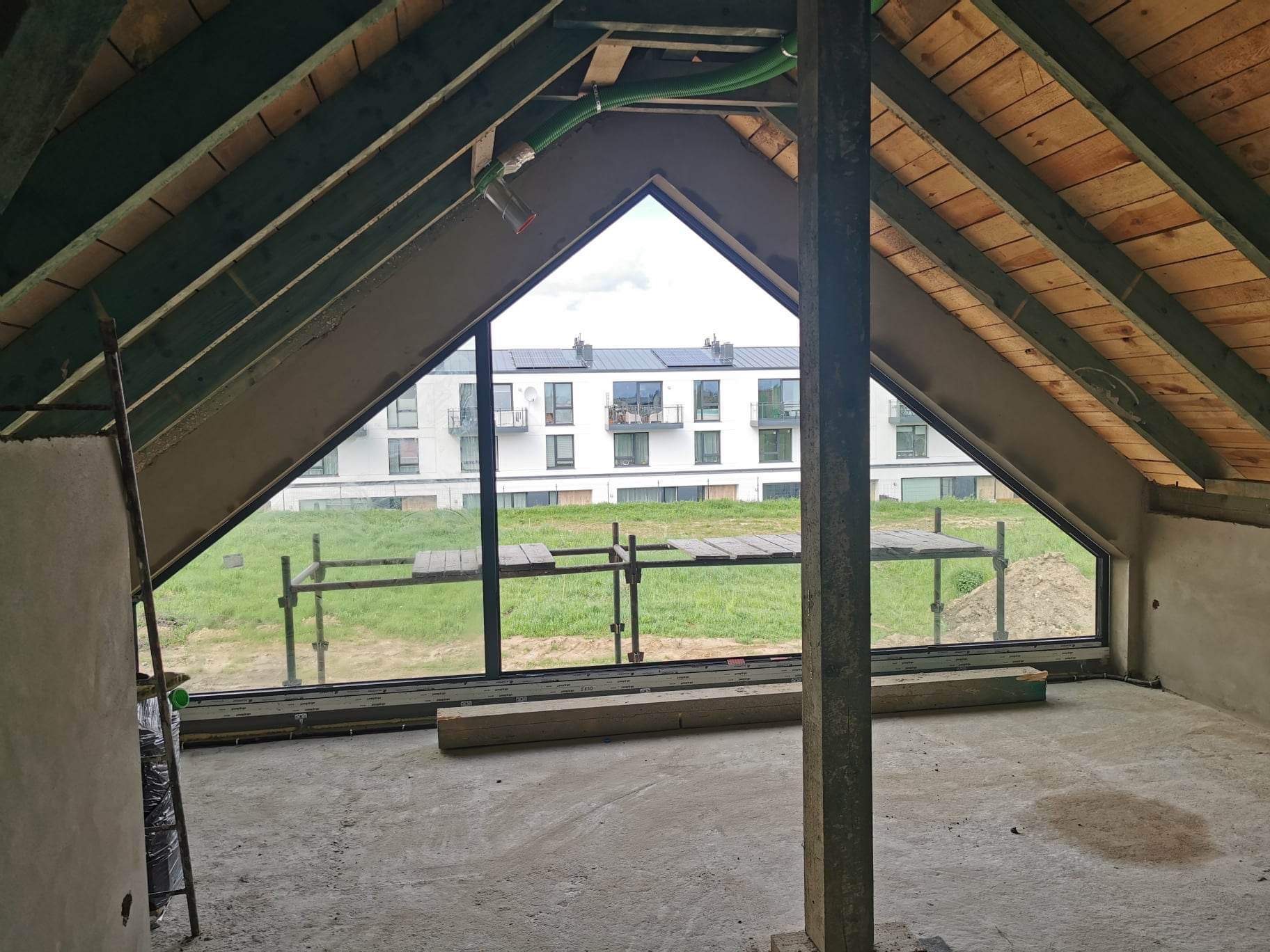 Poddasze w trakcie prac wykończeniowych, widoczne krokwie, okno z widokiem na budynek mieszkalny i zewnątrz, surowy beton na ścianach i podłodze, rusztowanie w tle