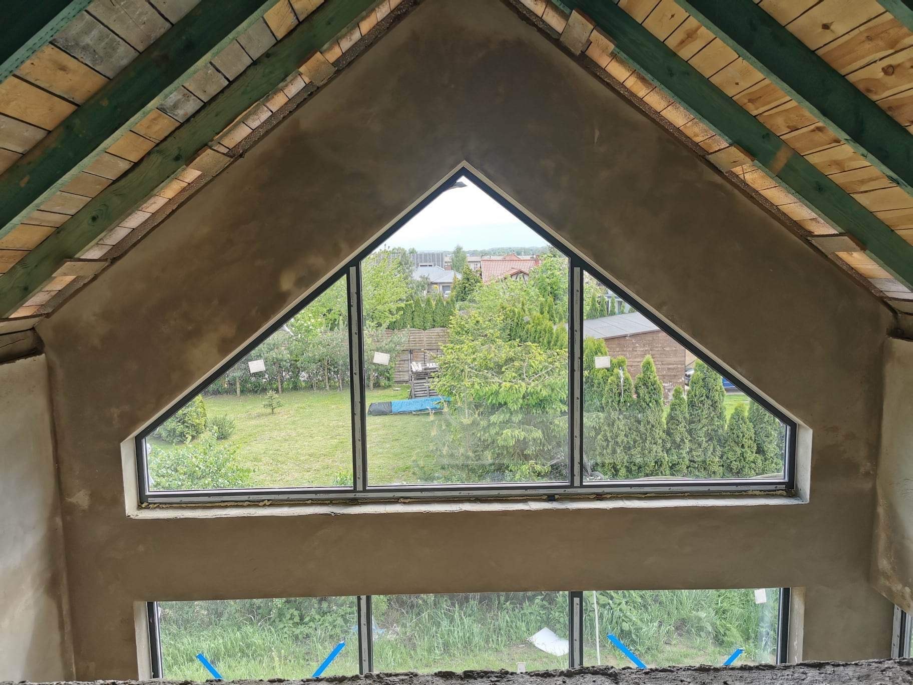Wnętrze poddasza z surowymi ścianami, widoczne okno trójkątne z widokiem na ogród, drewniana konstrukcja dachu z zielonymi belkami.