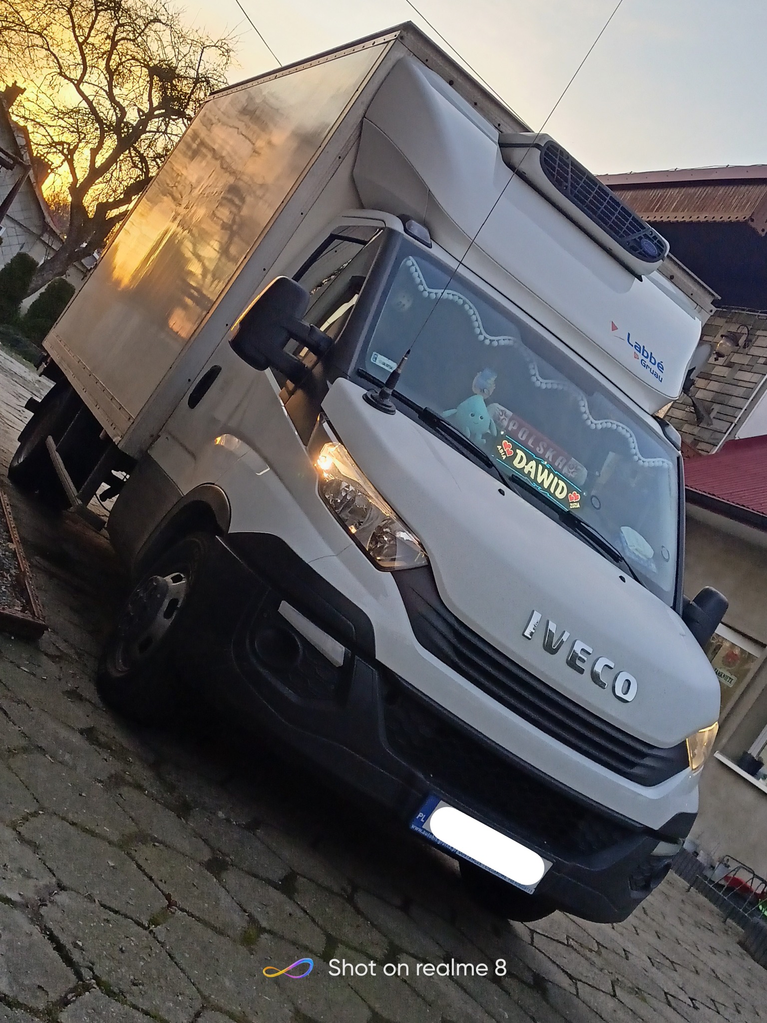 Biały samochód dostawczy Iveco z chłodnią, zaparkowany na brukowanej nawierzchni, z widocznym napisem 'DAWID' na desce rozdzielczej i pluszową maskotką w kabinie.