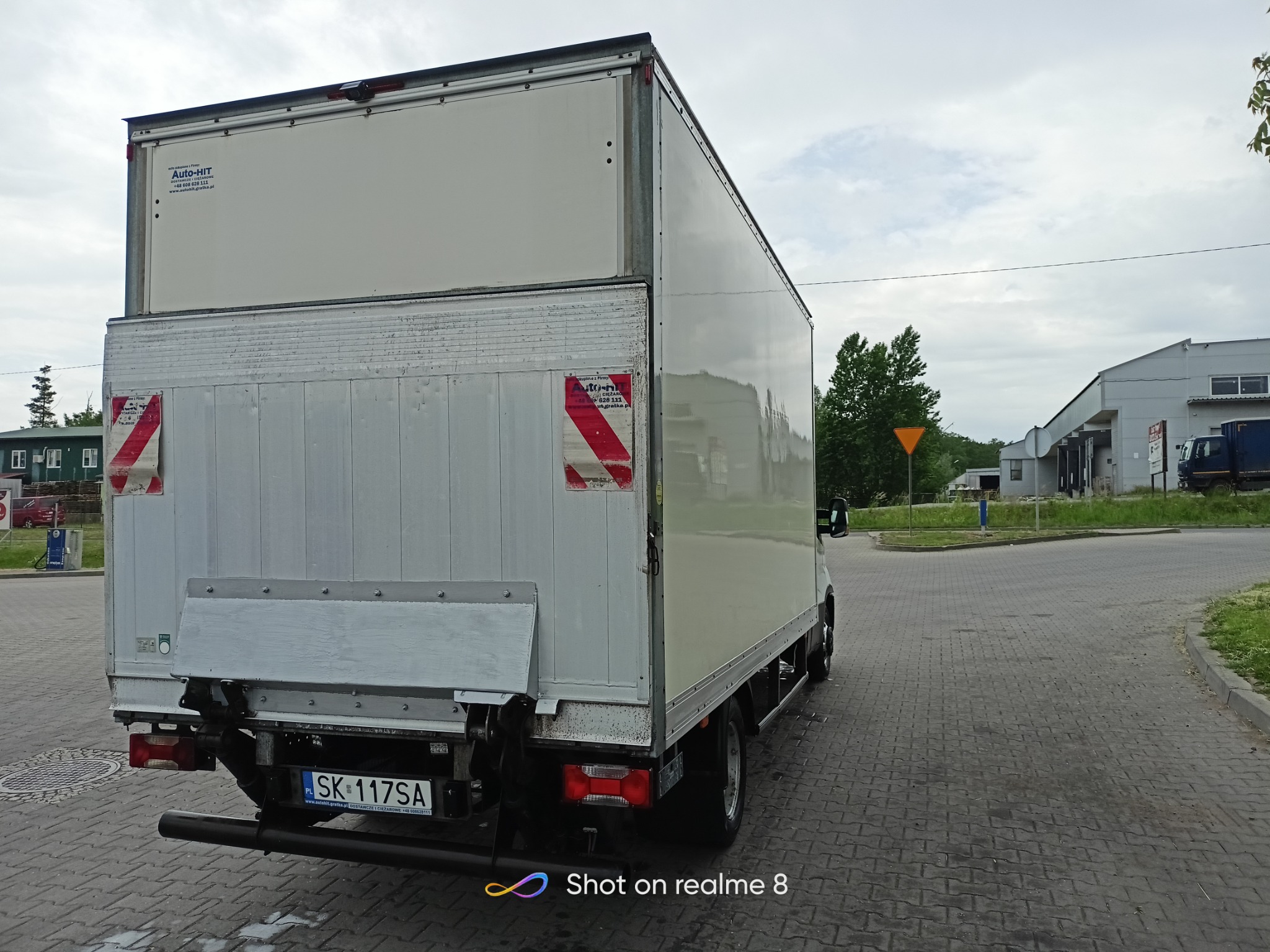 Tył białego samochodu dostawczego z windą załadowczą na parkingu z kostki brukowej, widoczne oznaczenia firmy Auto-HIT i tablica rejestracyjna SK 117SA.