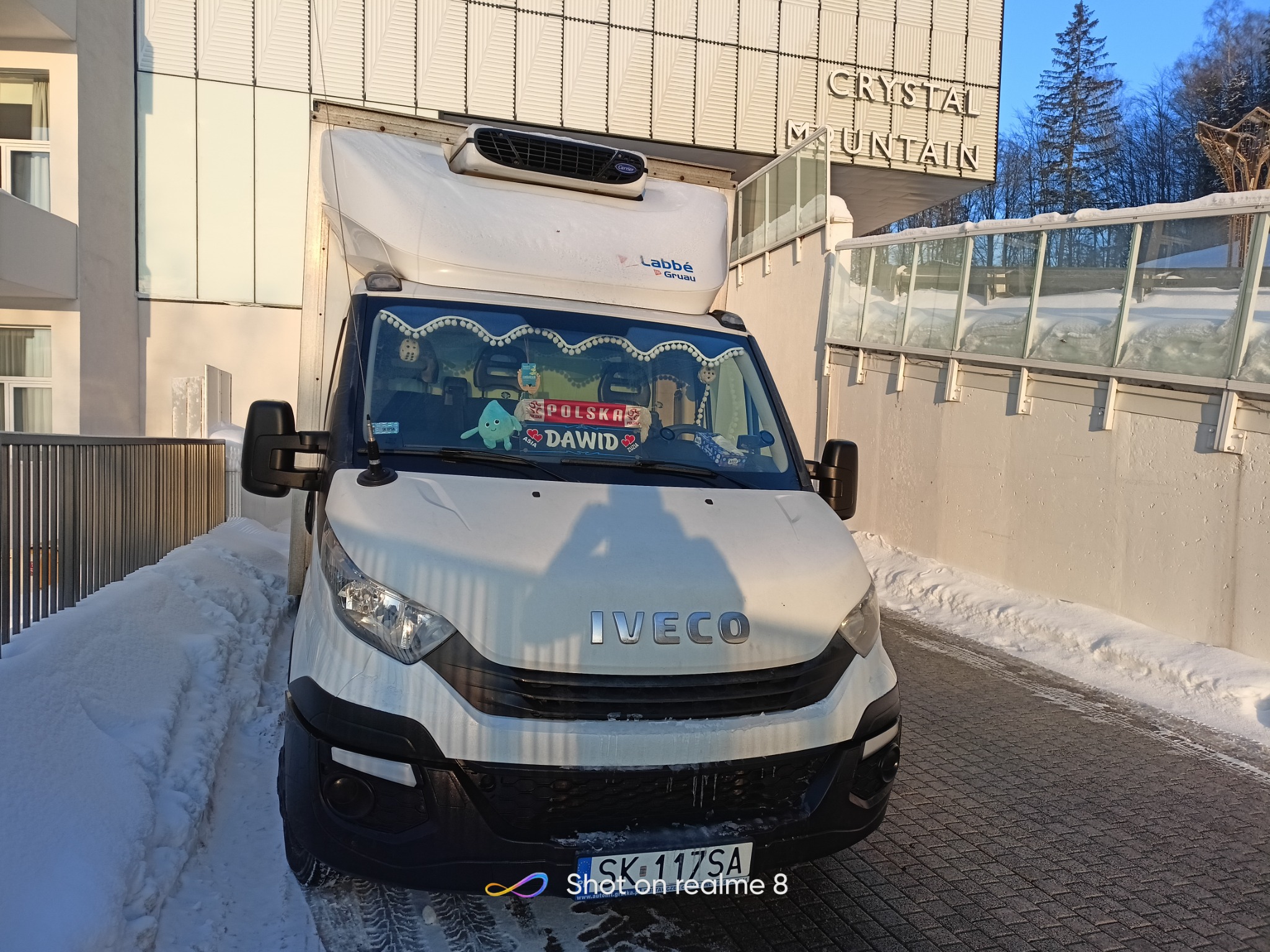 Biały samochód dostawczy Iveco z chłodnią Labbé-Gruau zaparkowany przed budynkiem Crystal Mountain w zimowej scenerii, ozdobiony pluszową zabawką i napisem 'Polska Dawid' na przedniej szybie.