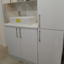 Biała szafka łazienkowa z umywalką nablatową i baterią, na tle płytki imitującej marmur oraz pralki.