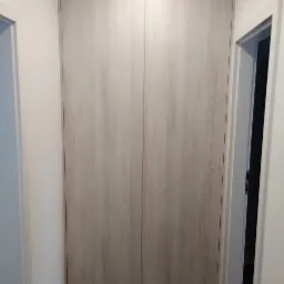 Zamknięte drzwi szafy wnękowej z jasnego drewna z widocznym usłojeniem, wkomponowane w białą ścianę, obok widoczne białe futryny drzwi.