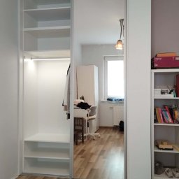 J&K Meble na Wymiar JANUSZ RYCHLICA - Biała szafa wnękowa z lustrzanymi drzwiami przesuwnymi, oświetleniem wewnętrznym i półkami, widoczna w pokoju z biurkiem i regałem.