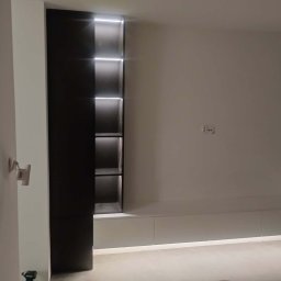 J&K Meble na Wymiar JANUSZ RYCHLICA - Minimalistyczna szafa wnękowa z podświetlanymi półkami i dolną zabudową w odcieniach bieli i czerni, wkomponowana w jasne wnętrze. Widoczny fragment podłogi i ściany.