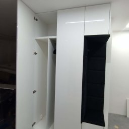 J&K Meble na Wymiar JANUSZ RYCHLICA - Biała szafa wnękowa z otwartymi drzwiami, widoczne półki i wieszaki. Obok szafy czarna, tapicerowana wnęka do siedzenia. Minimalistyczny design, nowoczesne oświetlenie.