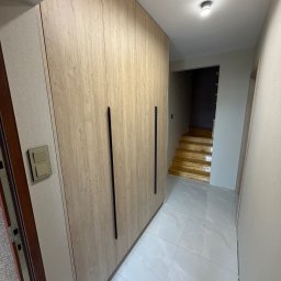 J&K Meble na Wymiar JANUSZ RYCHLICA - Wnętrze: zabudowa szafy wnękowej z drewnopodobnymi frontami i czarnymi uchwytami, widok na korytarz z białymi płytkami i schodami w tle.