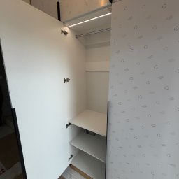 J&K Meble na Wymiar JANUSZ RYCHLICA - Otwarta, biała szafa wnękowa z oświetleniem LED i półkami, widok z bliska. Wnętrze wykończone płytą z subtelnym wzorem. Minimalistyczny design.