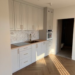 J&K Meble na Wymiar JANUSZ RYCHLICA - Nowoczesna kuchnia na wymiar z jasnoszarymi szafkami, drewnianym blatem, złotą baterią i uchwytami. Kwiatowy backsplash, drewniana podłoga w jodełkę, promienie słońca.