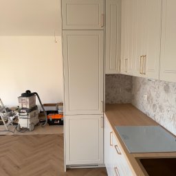 J&K Meble na Wymiar JANUSZ RYCHLICA - Delikatnie szare szafki kuchenne w stylu shaker z frezowanymi frontami i złotymi uchwytami. Drewniany blat, kwiecisty panel ścienny, płyta indukcyjna i wycięcie na zlew. W tle sprzęt budowlany.