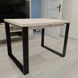 Jasny, kwadratowy stół z drewnianym blatem i czarnymi, metalowymi nogami w nowoczesnym wnętrzu.