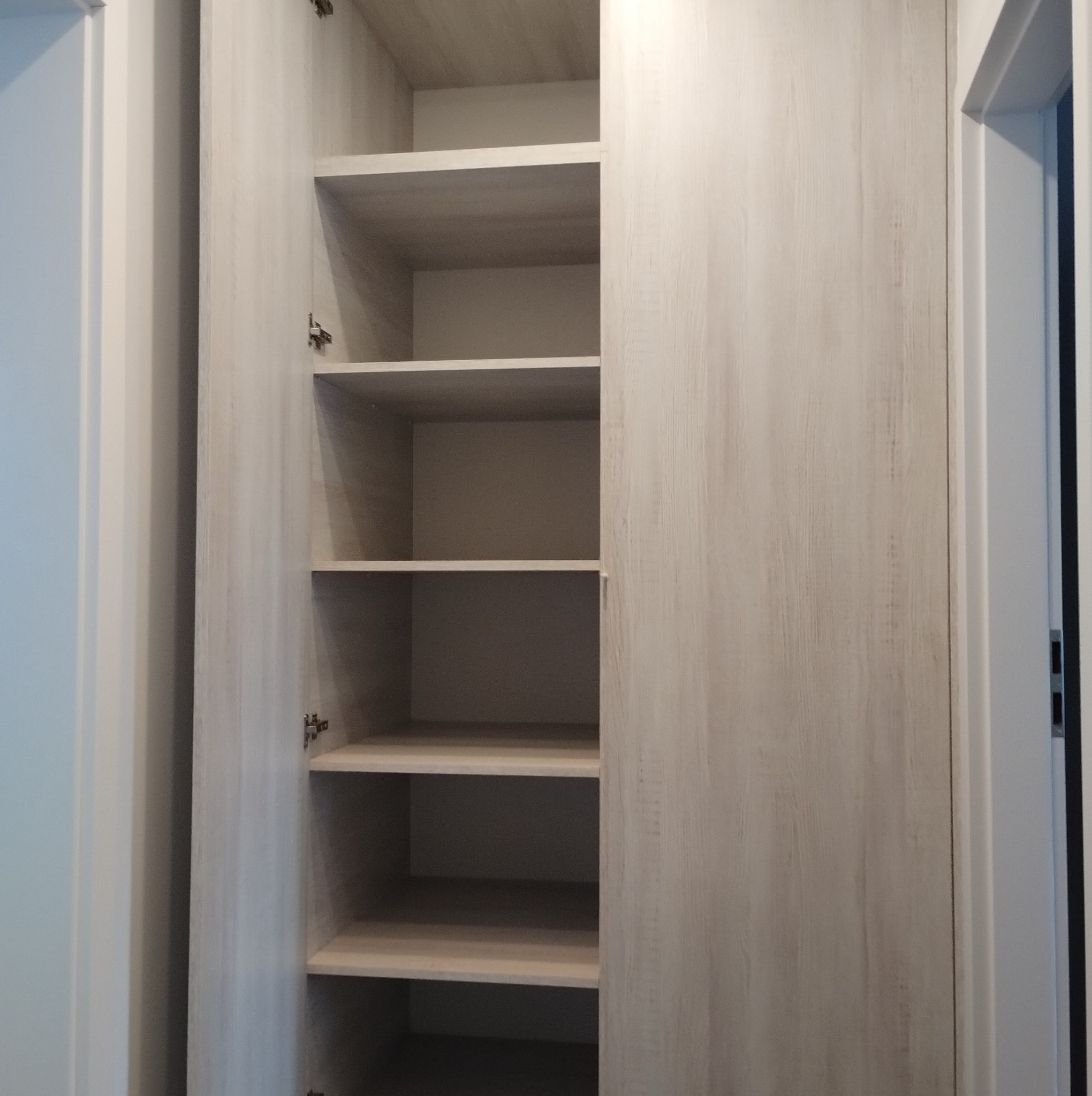 Otwarta szafa wnękowa z jasnego drewna z półkami, widoczne zawiasy, wnętrze puste.