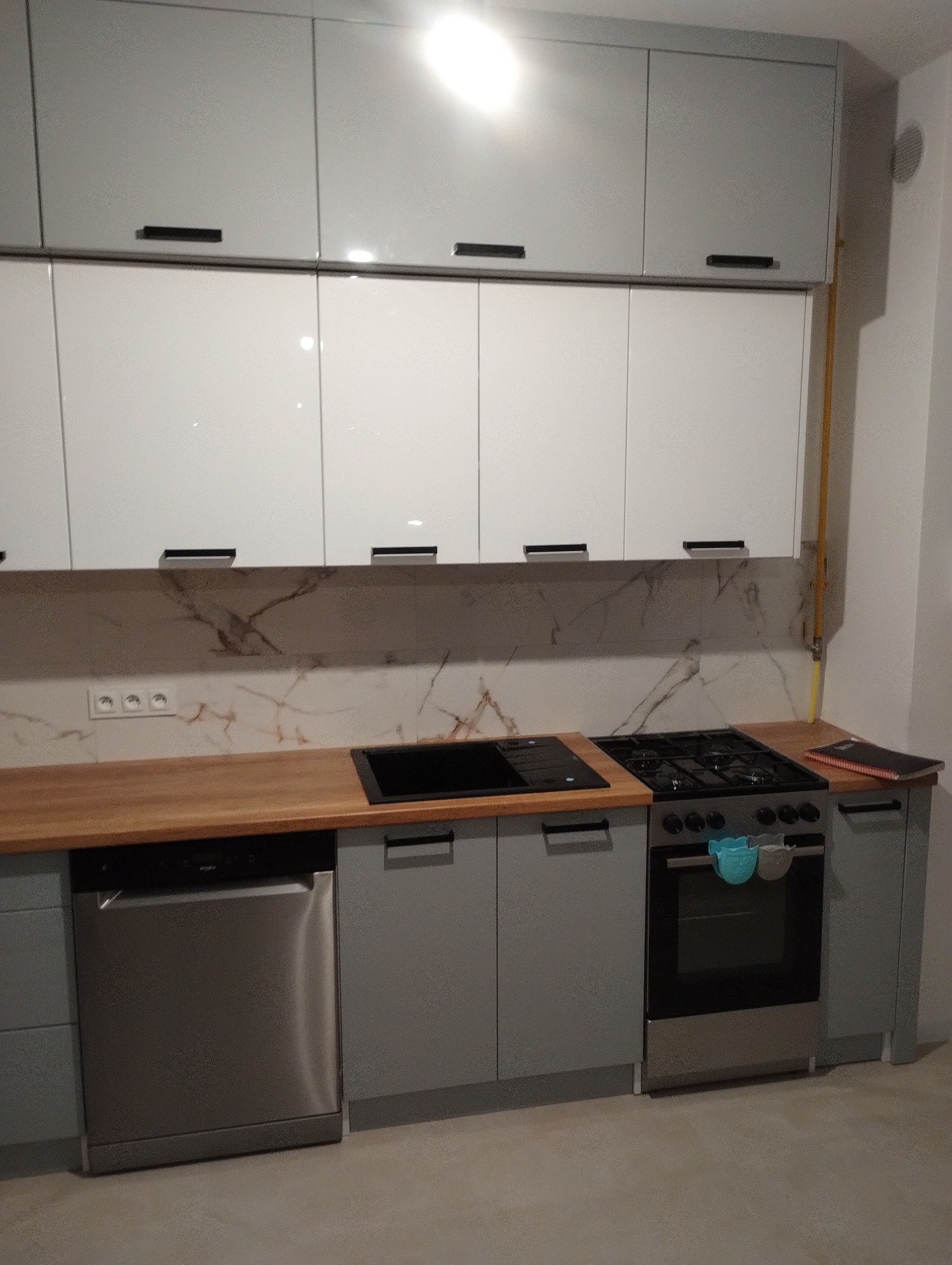 Szare i białe szafki kuchenne z drewnianym blatem, czarnym zlewem i płytą gazową; na piekarniku wiszą ściereczki.