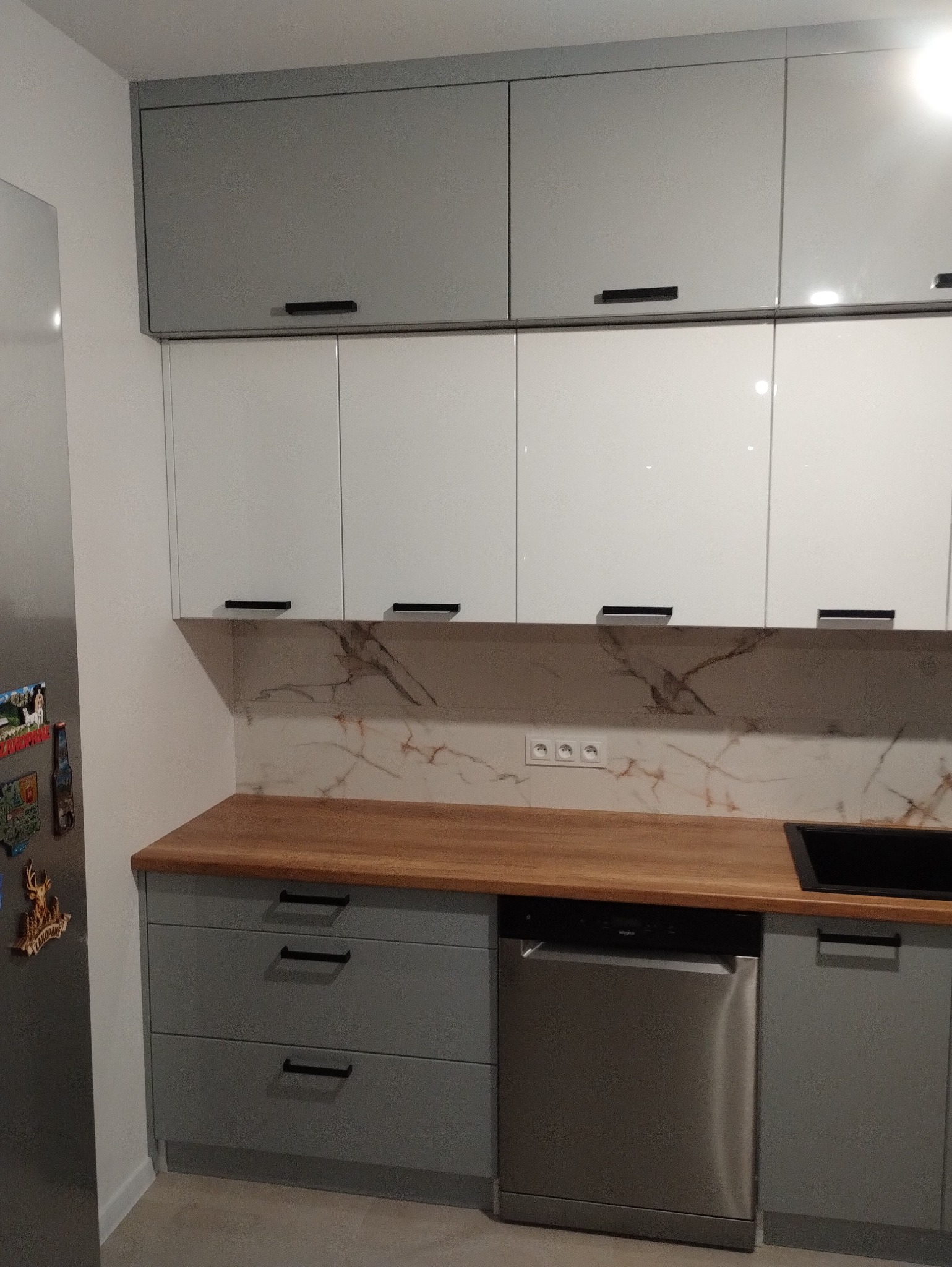 Nowoczesna kuchnia z szarymi i białymi szafkami, drewnianym blatem i marmurowym backsplashiem, uzupełniona o czarne uchwyty i srebrną zmywarkę.