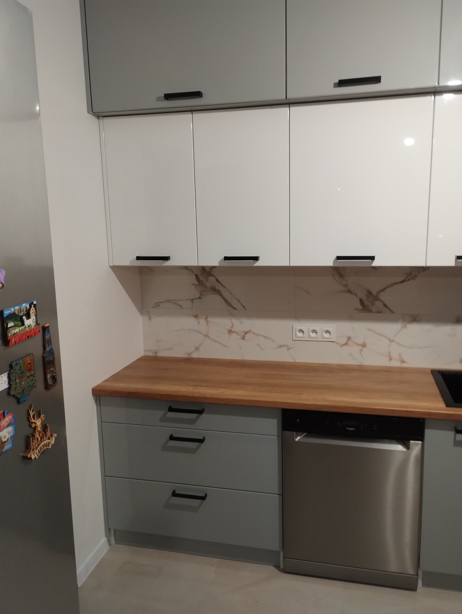 Nowoczesna kuchnia z szarymi dolnymi szafkami z trzema szufladami, drewnianym blatem, zmywarką i białymi górnymi szafkami z szarymi szafkami nad nimi, marmurowy panel nad blatem, magnesy na lodówce.