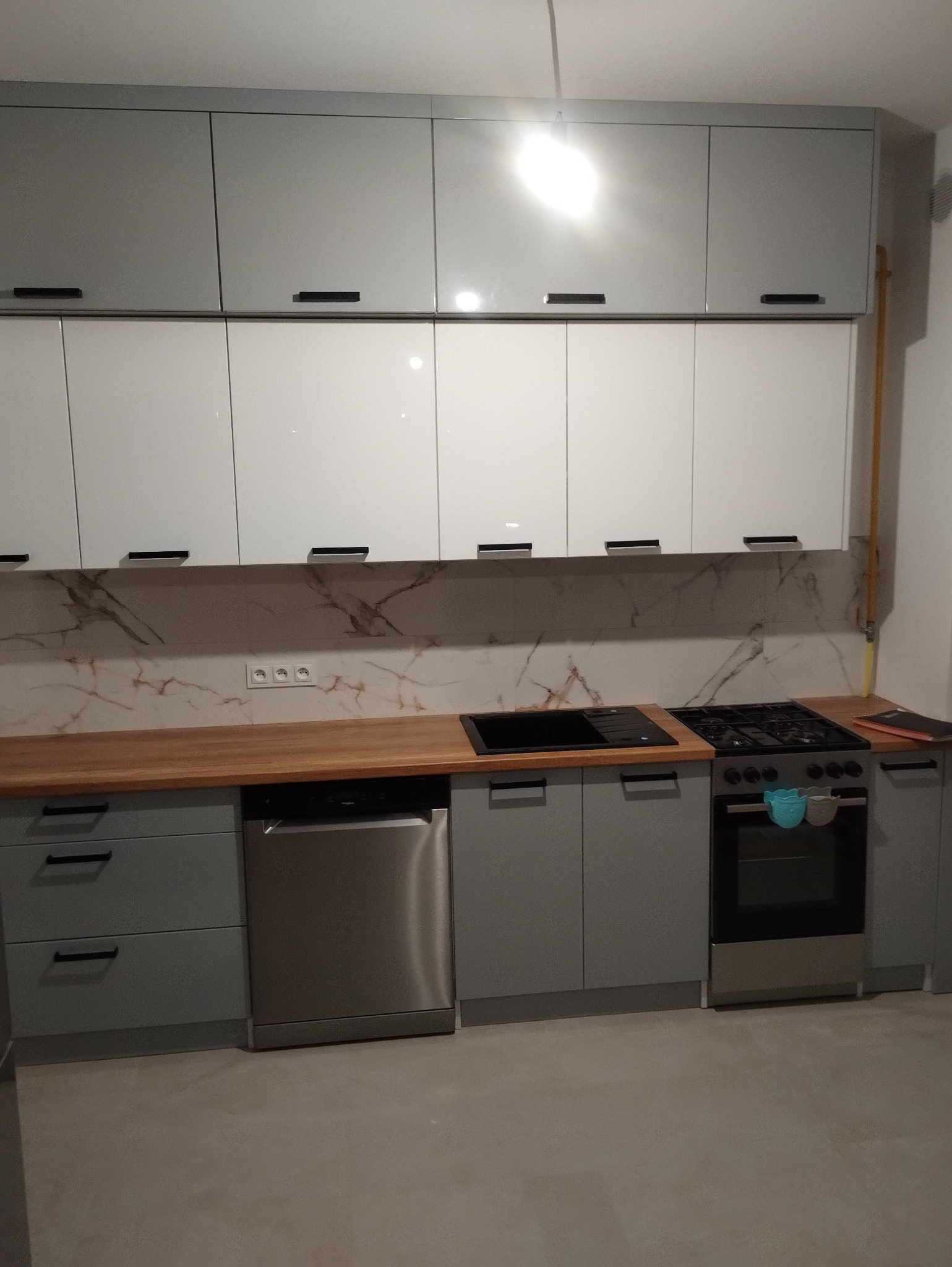 Szaro-biała zabudowa kuchenna z drewnianym blatem, marmurowym panelem nad blatem, czarnym zlewem i płytą gazową; na piekarniku wiszą ściereczki.