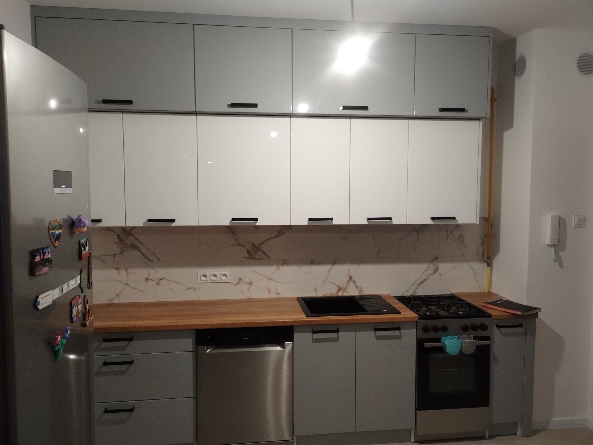 Nowoczesna kuchnia w odcieniach szarości i bieli z drewnianym blatem, marmurowym backsplashiem i lodówką z magnesami.