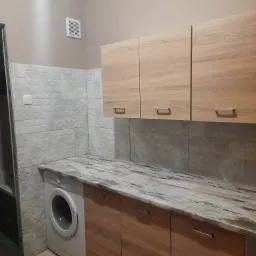 Kuchnia z pralką, jasne drewniane szafki górne i dolne, blat imitujący marmur, ściana wyłożona białymi płytkami z geometrycznym wzorem, beżowa ściana górna z kratką wentylacyjną.