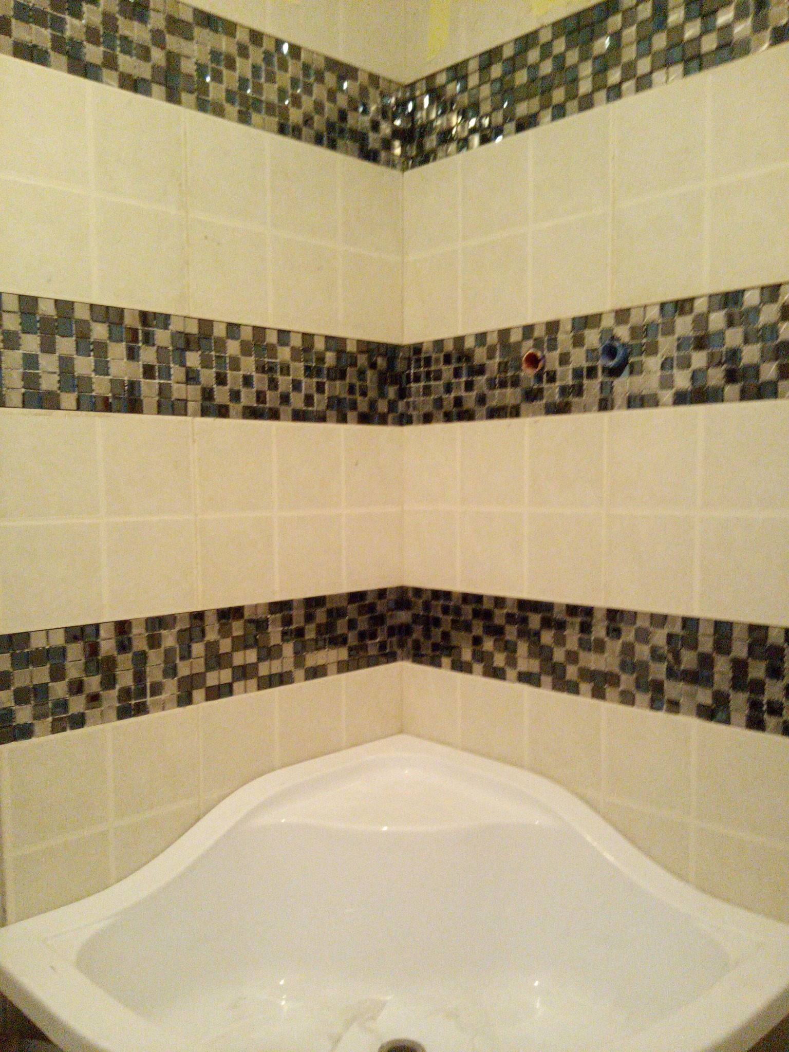 Wykończona ściana kabiny prysznicowej z białą armaturą, zdobiona pasami mozaiki w odcieniach szarości i błękitu, z widocznymi przyłączami wodnymi.