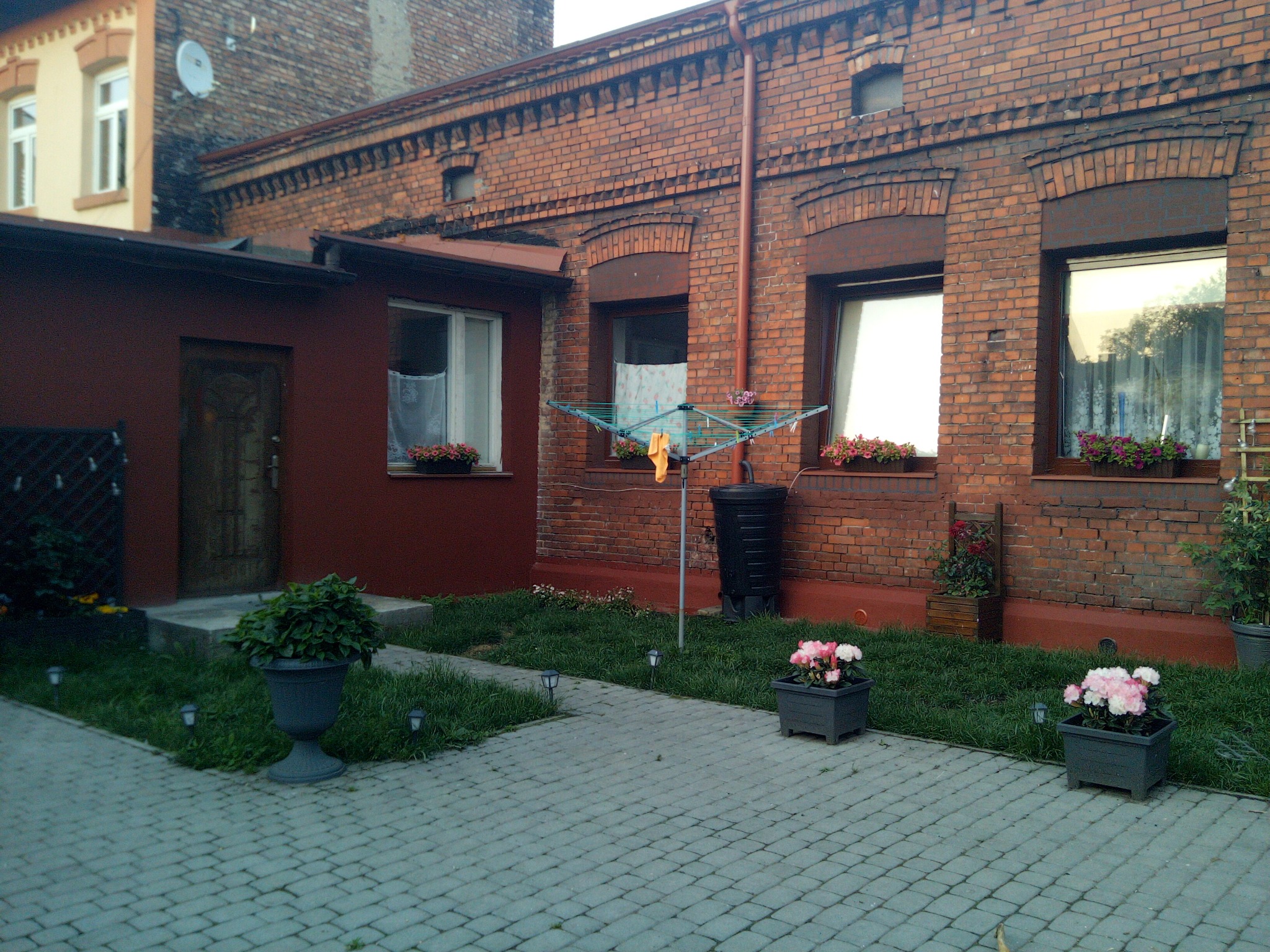Dziedziniec kamienicy z cegły z odnowioną elewacją, kwiatami w donicach na parapetach i chodnikiem z kostki brukowej