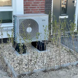 Szara pompa ciepła zamontowana na zewnątrz budynku, otoczona kamieniami i niskimi krzewami z młodymi listkami, widoczna elewacja z czerwonej cegły i szare drzwi wejściowe.