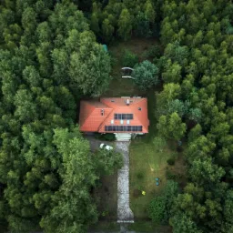 Widok z drona na dom z czerwoną dachówką, otoczony gęstym lasem; na dachu widoczne panele fotowoltaiczne, a obok domu zaparkowany biały samochód.