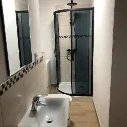 Nowoczesna łazienka po remoncie: kabina prysznicowa z czarnymi ramami i deszczownicą, umywalka z chromowaną baterią, beżowe płytki na ścianach i podłodze, pralka schowana w rogu.