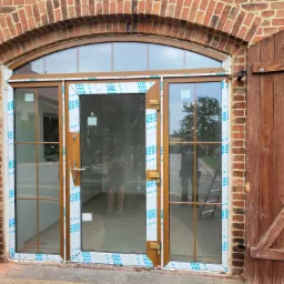 Nowe, dwuskrzydłowe okno z elementami drzwiowymi, z brązowymi szprosami i ramą, w trakcie montażu, zabezpieczone folią ochronną, widoczne ceglane obramowanie i drewniane okiennice.