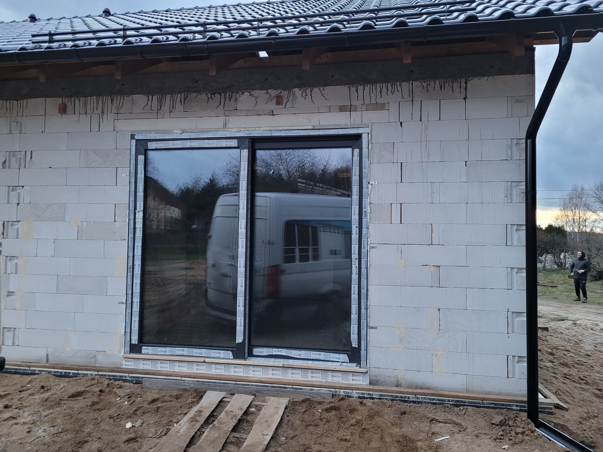 Nowo zamontowane, przesuwne okno w surowym stanie, widoczne zabezpieczenia folią, odbicie samochodu w szybie, ściana z betonu komórkowego w trakcie budowy.