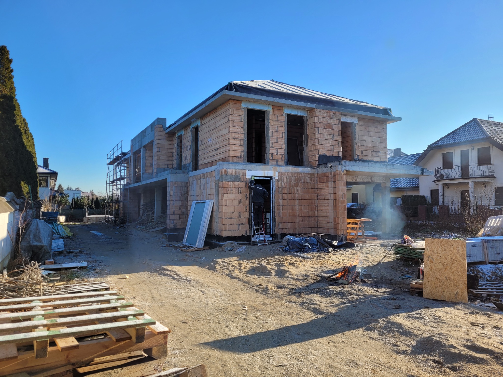 Surowy stan domu w trakcie budowy z widoczną stertą palet, ogniskiem i oknem opartym o ścianę, w tle inne zabudowania.