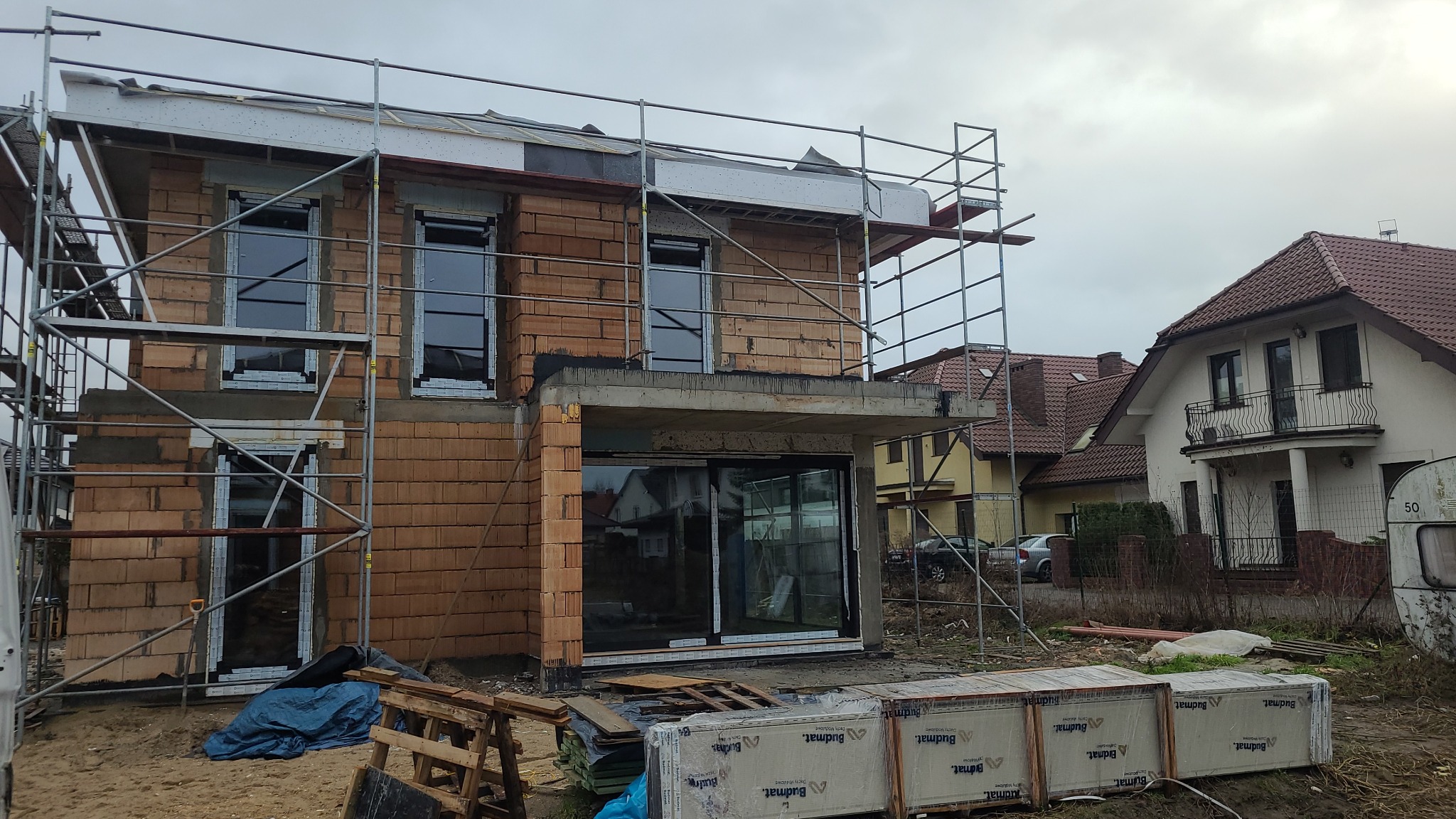 Nowy dom w trakcie budowy, widoczne rusztowania, wstawione okna PCV, materiały budowlane Budmat na ziemi, pochmurne niebo.