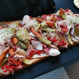 Drewniana deska obficie załadowana wędlinami, serami, ogórkami kiszonymi, kiełbasami, czerwoną porzeczką i świeżymi kiełkami, całość tworzy apetyczną kompozycję.