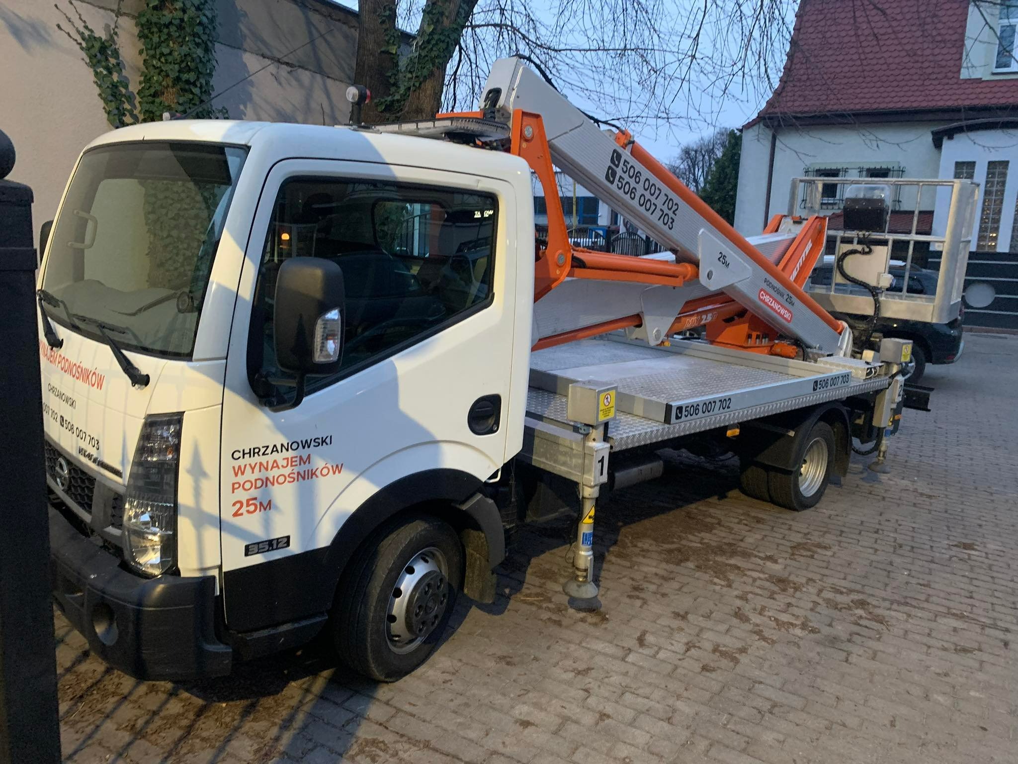 Samochód z podnośnikiem koszowym na parkingu z kostki brukowej, widoczne numery kontaktowe firmy na ramieniu podnośnika i kabinie pojazdu.
